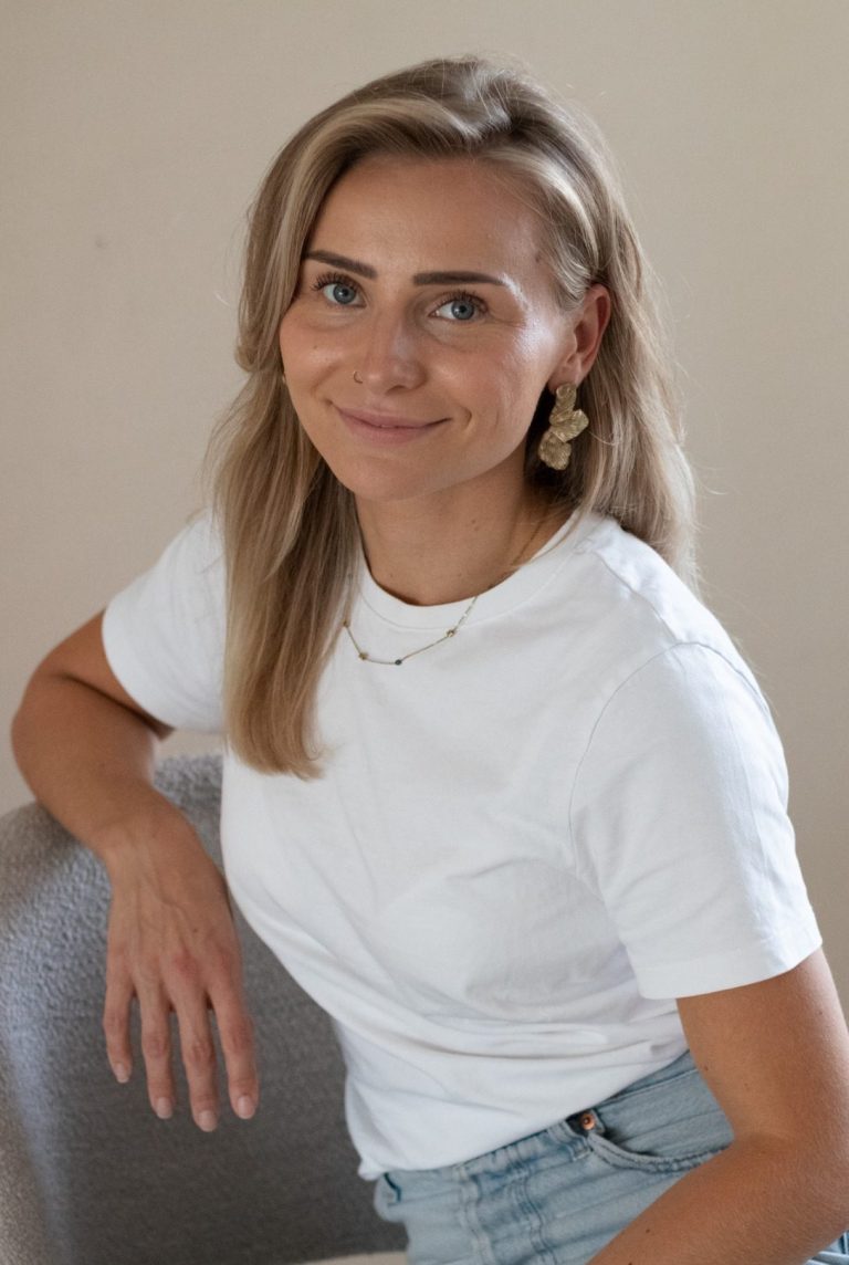 Jonge vrouw met blond haar, in een wit shirt, glimlachend en zittend op een stoel.