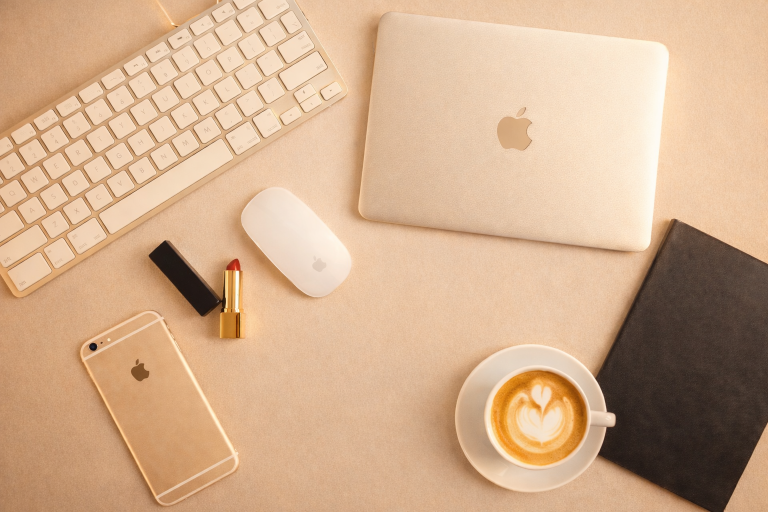 Een gouden laptop, toetsenbord, muis, iPhone, lippenstift en een kopje koffie op een bureau.