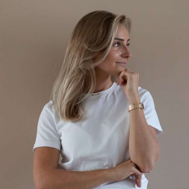 Vrouw met lang, blond haar, in een wit T-shirt, staand met een nadenkende uitdrukking.